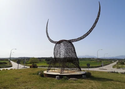 "Imponente escultura de hierro del Cap de Dimoni en la entrada de Santa Margalida, Mallorca." "Monumental obra del Cap de Dimoni, emblema de la tradición y la artesanía de Santa Margalida." "La Escultura del Cap de Dimoni en Santa Margalida, un punto de encuentro cultural en Mallorca." "Vista de la escultura del Cap de Dimoni en Santa Margalida, reflejo de las fiestas de la Beata." "El Cap de Dimoni de Santa Margalida, un hito artístico forjado en hierro en el corazón de Mallorca."